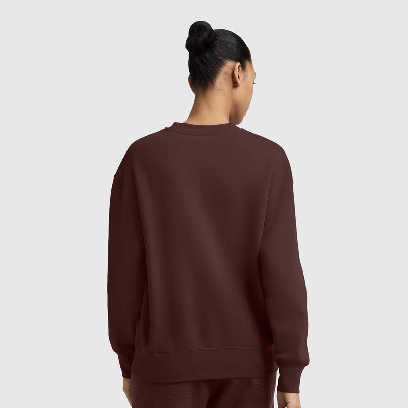 A person with dark hair in a bun is shown from the back, wearing a loose maroon Champion Powerblend Crewneck Sweatshirt with an embroidered script logo and matching pants, standing against a plain light gray background.