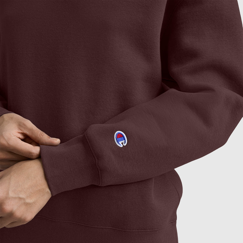 A person wearing a maroon Champion Powerblend Crewneck Sweatshirt, Embroidered Script Logo, with the signature Champion logo on the sleeve, adjusts the cuff with their hand. The background is plain and light-colored.