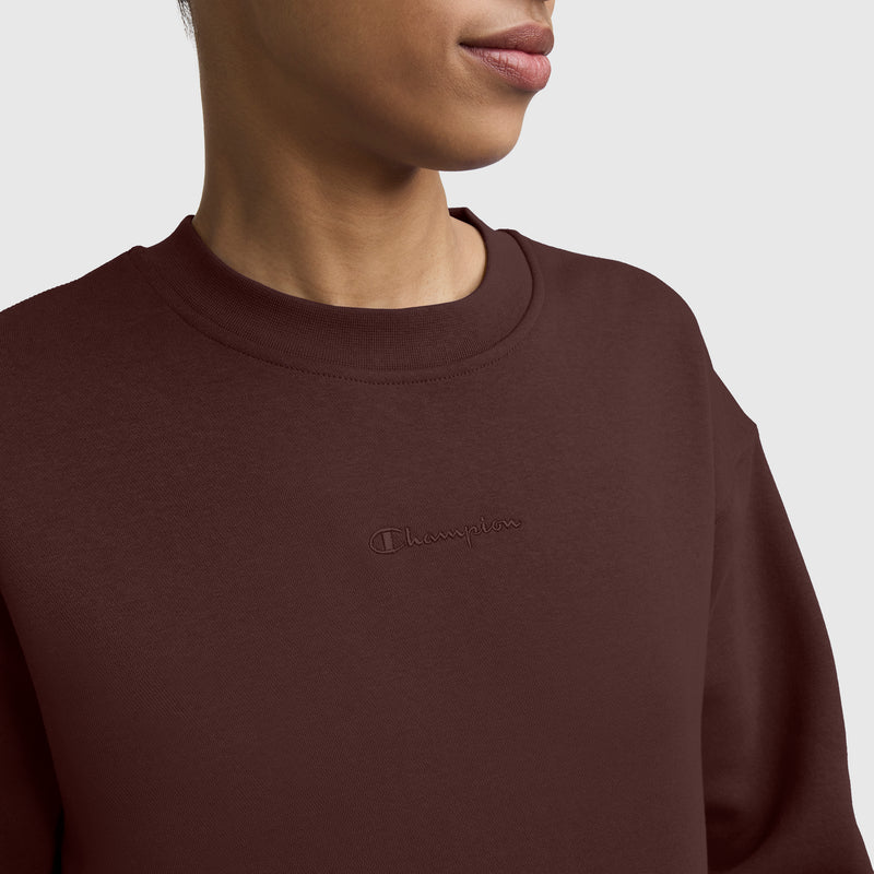A person is wearing the Champion Powerblend Crewneck Sweatshirt, Embroidered Script Logo, in dark brown. The focus is on the upper torso and part of the face against a plain light gray background. This classic sweatshirt for women features soft Powerblend fleece for all-day comfort and style, with the Champion logo embroidered at the center.