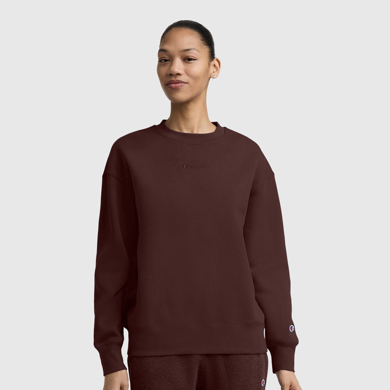 A person wearing a burgundy Champion Powerblend Crewneck Sweatshirt with embroidered script logo and matching pants stands against a plain gray background, smiling slightly.