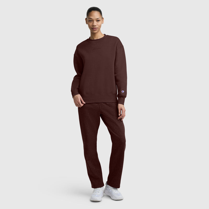 A person stands against a plain background wearing a maroon Champion Powerblend Crewneck Sweatshirt with an Embroidered Script Logo, paired with pants and white sneakers, looking at the camera with one hand in their pocket.