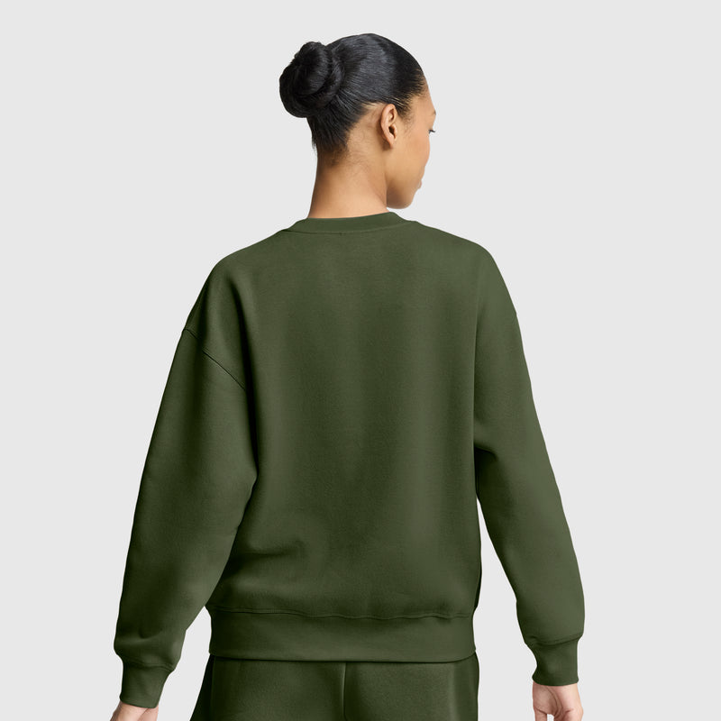 A woman with her hair in a bun, dressed in an olive green Champion Powerblend Crewneck Sweatshirt with an embroidered script logo and matching pants, stands with her back to the camera against a plain light gray background.