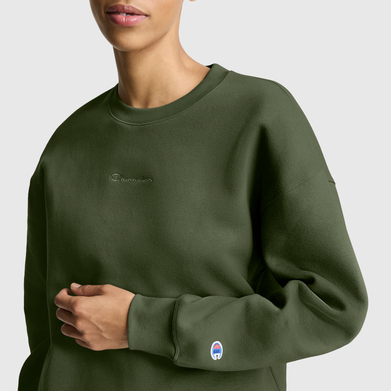 A woman is shown from shoulders to waist against a plain background wearing the Champion Powerblend Crewneck Sweatshirt in olive green, made with soft fleece and featuring small embroidered script logos on the chest and sleeve.