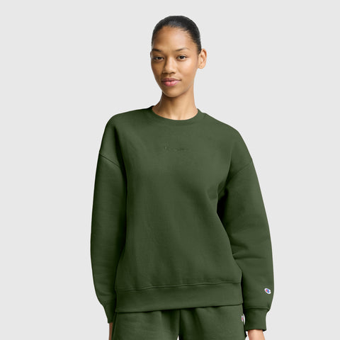 A woman dressed in an oversized dark green sweatshirt and matching pants stands against a plain light gray background, gazing at the camera with a neutral expression. Her relaxed outfit captures the essence of women’s fashion sweatshirts, similar to the Champion Powerblend Crewneck Sweatshirt with Embroidered Script Logo, renowned for its soft fleece comfort.