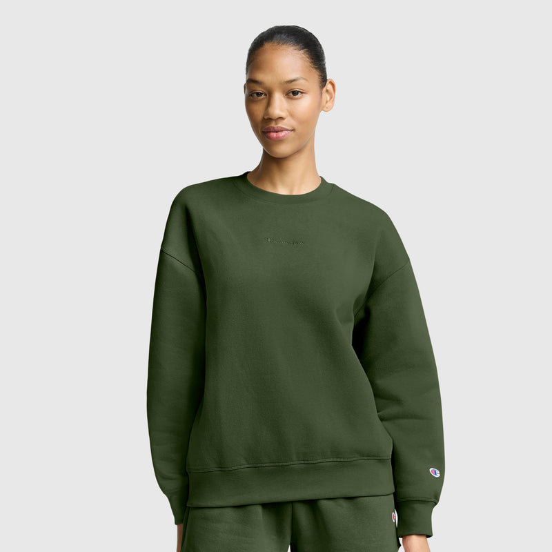 A woman dressed in an oversized dark green sweatshirt and matching pants stands against a plain light gray background, gazing at the camera with a neutral expression. Her relaxed outfit captures the essence of women’s fashion sweatshirts, similar to the Champion Powerblend Crewneck Sweatshirt with Embroidered Script Logo, renowned for its soft fleece comfort.