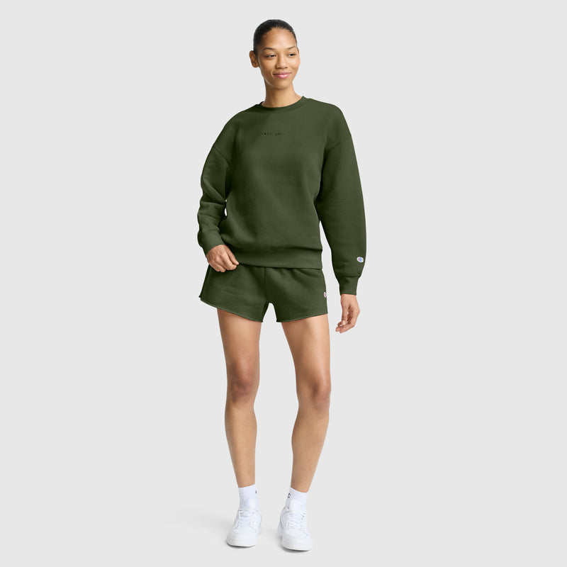 A woman stands against a plain background wearing a Champion Powerblend Crewneck Sweatshirt with an embroidered script logo in olive green, paired with matching shorts and white sneakers. Smiling with one hand in her pocket, she effortlessly showcases the latest in women’s fashion featuring Champion’s cozy Powerblend fleece.