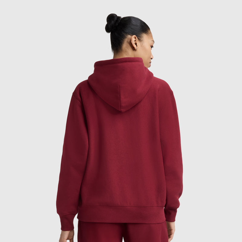 A person with dark hair in a bun is shown from the back, wearing a matching maroon Champion Essentials Reverse Weave Hoodie Sweatshirt and sweatpants against a plain light gray background. The hoodie features an embroidered C patch.