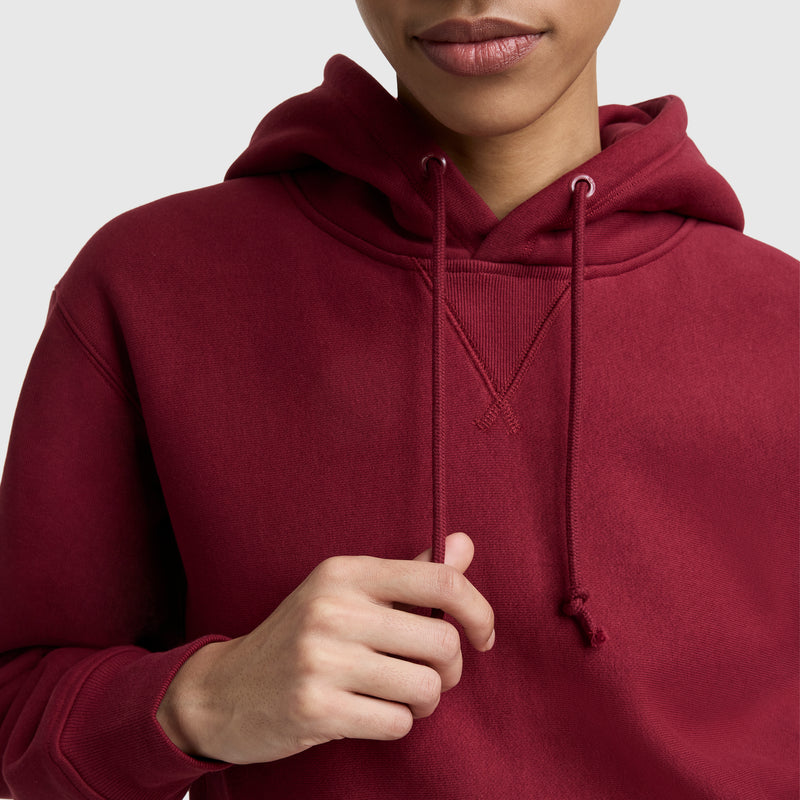 A person in a maroon Champion Essentials Reverse Weave Hoodie Sweatshirt is shown from shoulders to mouth, gently holding one hoodie drawstring with their right hand. The background is plain and light-colored.