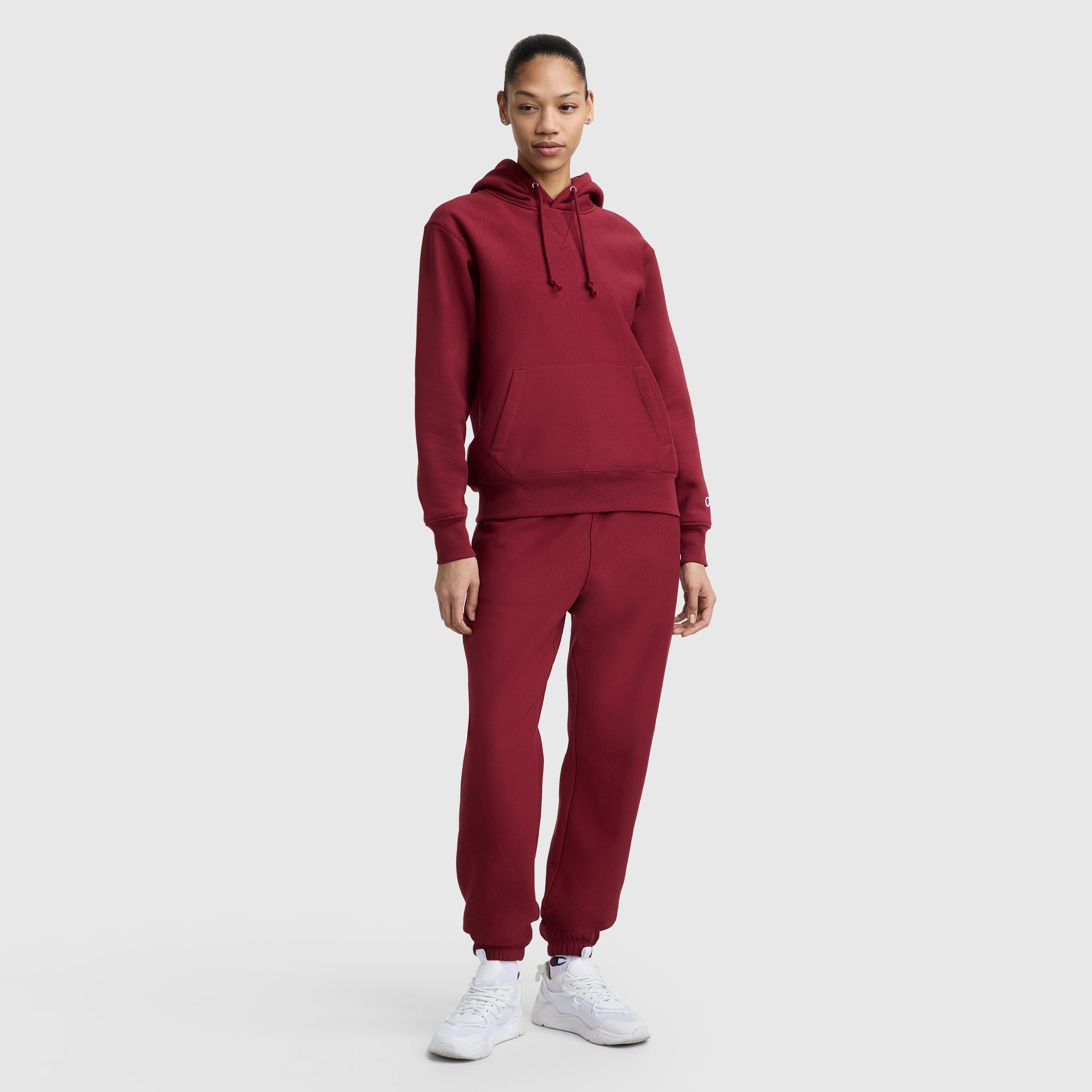 A person stands against a plain background wearing a matching maroon Champion Essentials Reverse Weave Hoodie Sweatshirt and sweatpants set with white sneakers. The heavyweight fleece hoodie features an embroidered C patch. They appear relaxed and neutral.