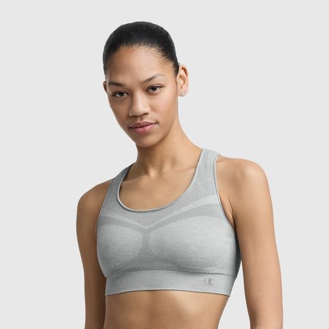 A woman with dark hair pulled back wears the Champion Seamless Racerback Sports Bra in light gray and small stud earrings, standing against a plain light background.