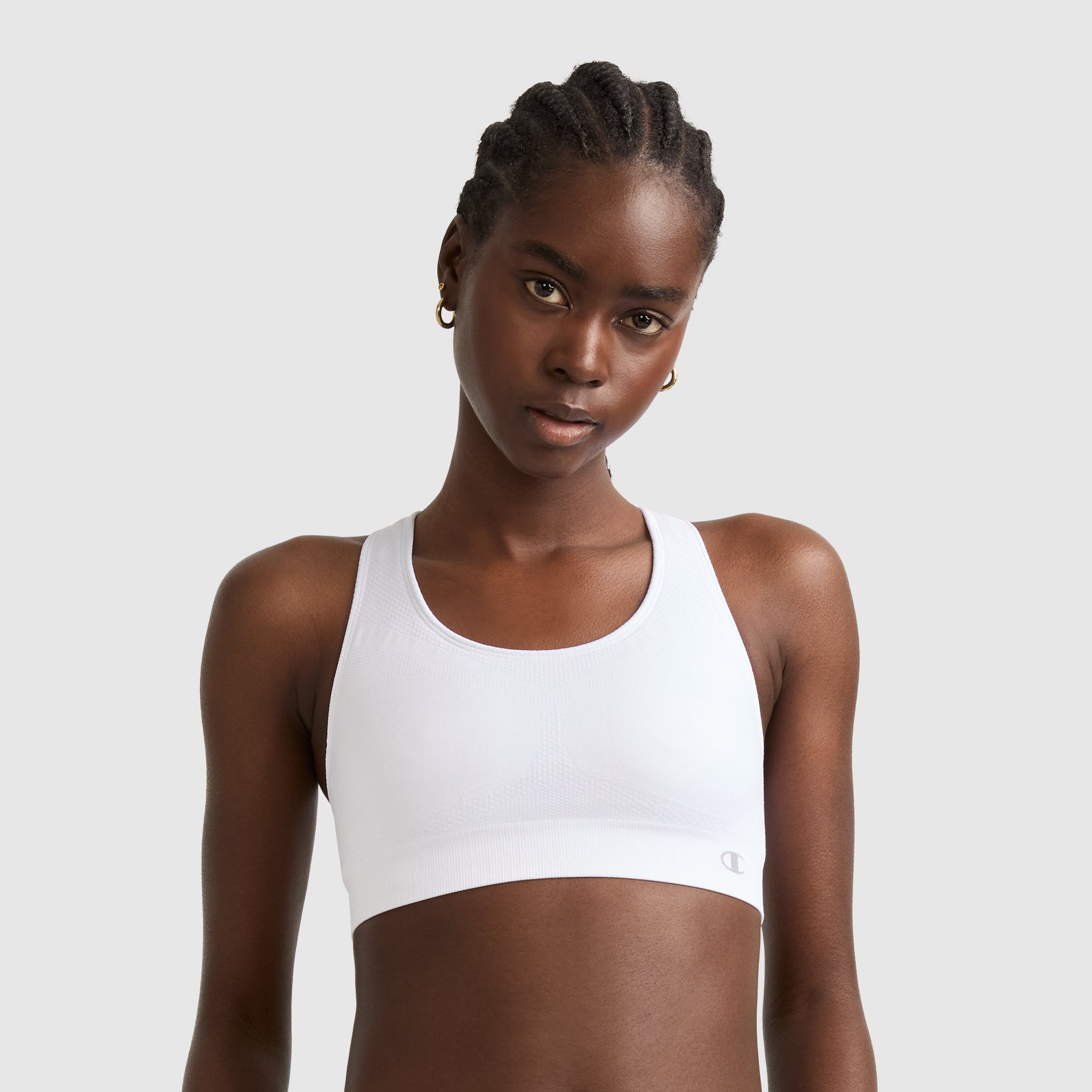 A woman with braided hair models the Champion Seamless Racerback Sports Bra in white with small gold hoop earrings, standing against a plain light gray background.