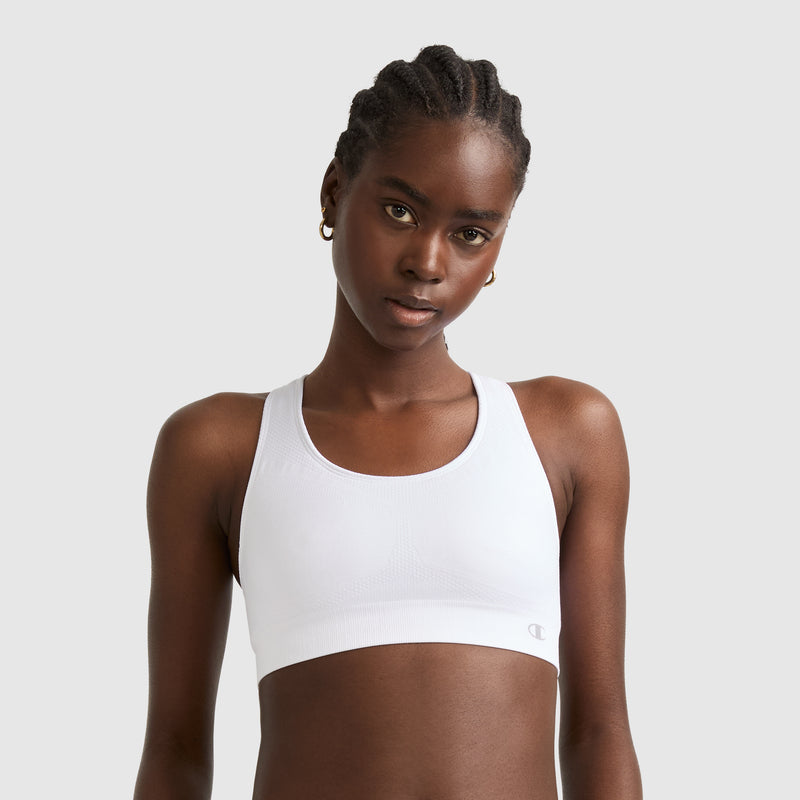 A woman with braided hair models the Champion Seamless Racerback Sports Bra in white with small gold hoop earrings, standing against a plain light gray background.