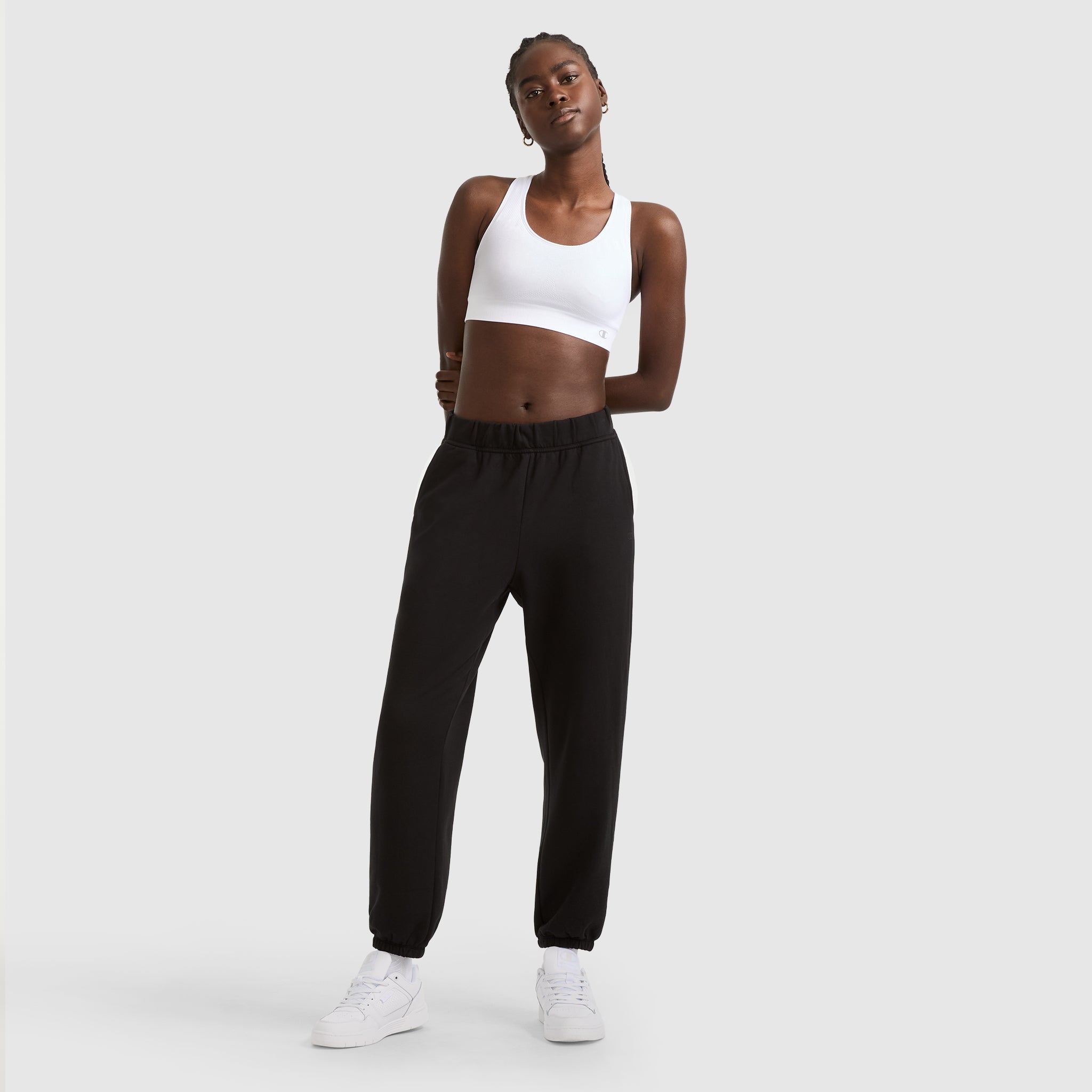 A person stands against a light gray background, wearing the Champion Seamless Racerback Sports Bra in white, paired with black jogger pants and white sneakers, hands behind their back and looking confidently ahead.