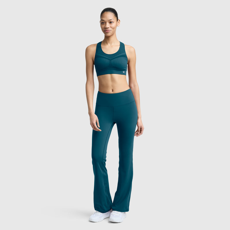 A woman stands against a plain background, wearing the Champion Seamless Racerback Sports Bra in teal, paired with matching high-waisted flared leggings and white sneakers. She looks forward with a relaxed expression and one hand by her side.