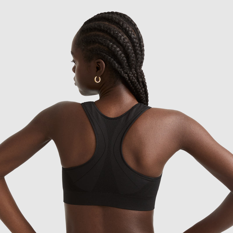 A woman with long, braided hair stands with her back to the camera, wearing a black racerback sports bra and gold hoop earrings, against a plain light gray background.