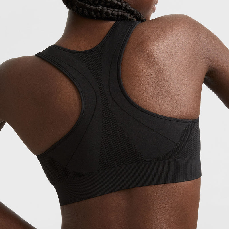 A woman with braided hair is shown from the back wearing a black racerback sports bra with textured panels and seamless detailing.