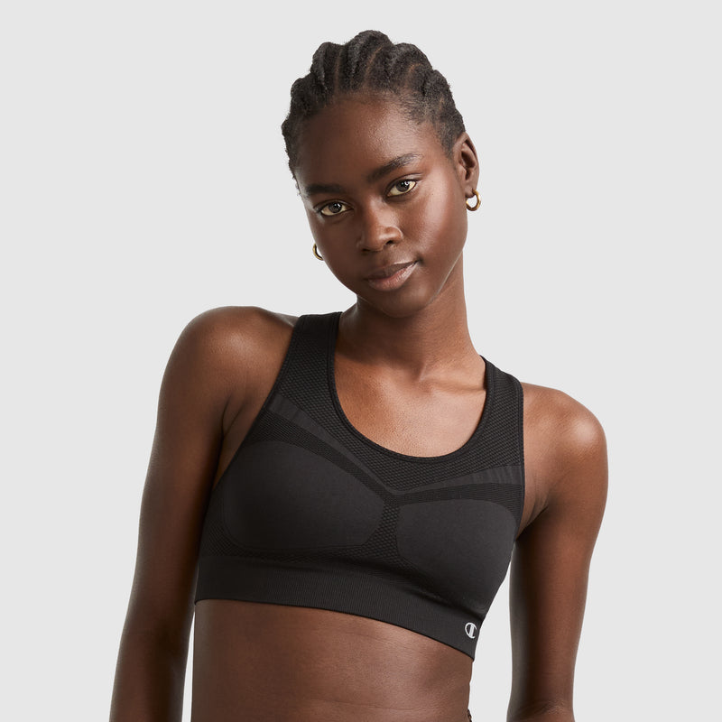 A woman with braided hair wears a black sports bra and gold hoop earrings, standing against a plain light gray background, looking into the camera with a slight smile.