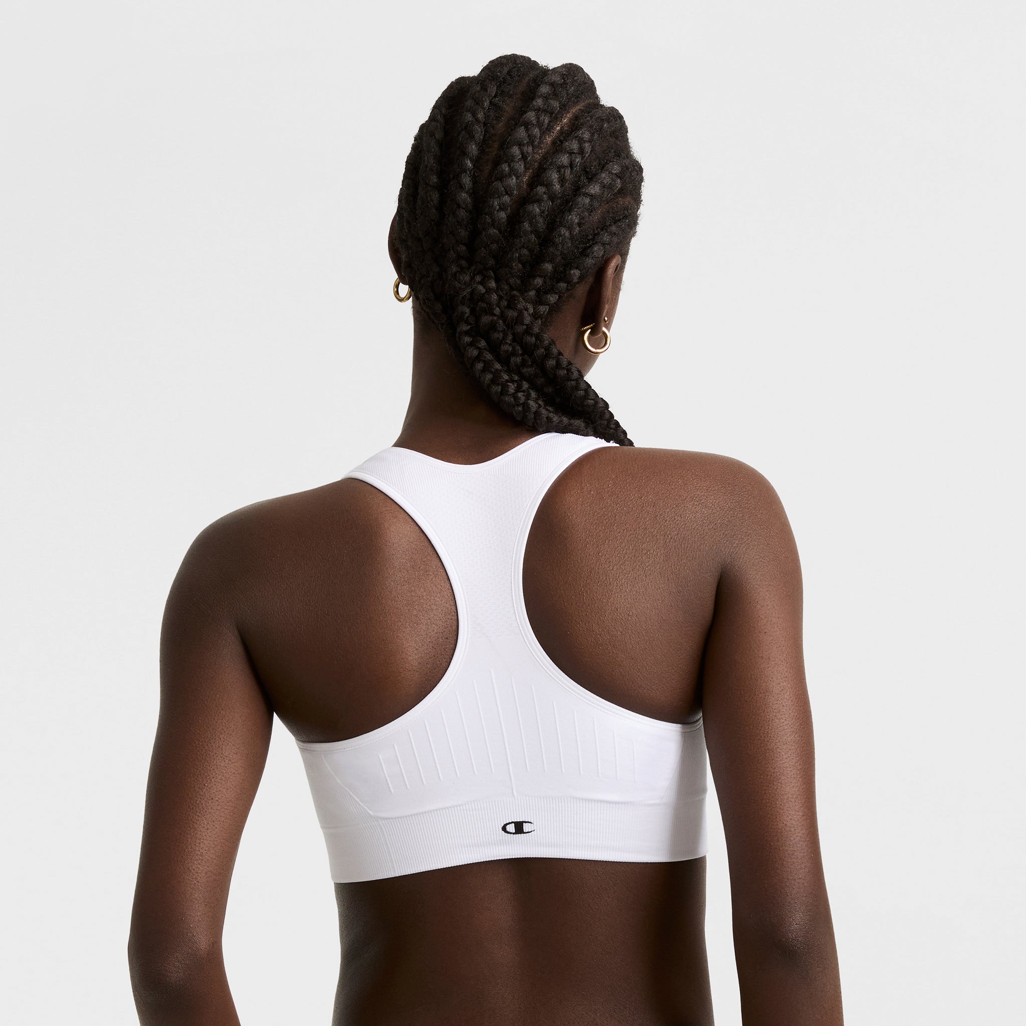 A woman with braided hair and small hoop earrings models the Champion Infinity Racerback Sports Bra, shown from behind against a plain light background.