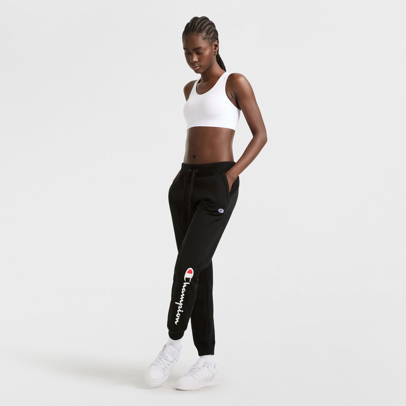 A woman with braided hair wears a white Champion Infinity Racerback Sports Bra and black Champion sweatpants, standing with hands in her pockets against a plain white background. She finishes the look with white sneakers.