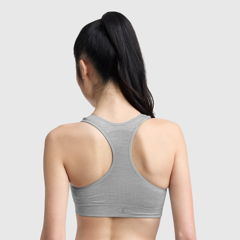 A woman with long dark hair in a high ponytail is shown from the back, wearing the Champion Infinity Racerback Sports Bra—a light gray wireless style—against a plain light gray background.