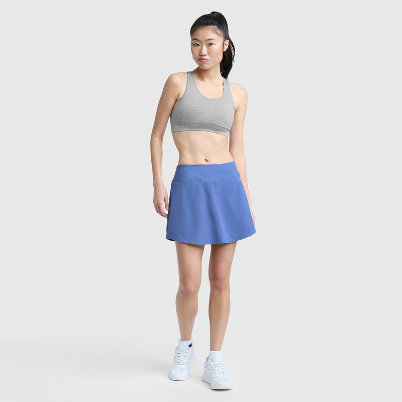 A woman stands against a plain background wearing a Champion Infinity Racerback Sports Bra, blue athletic skirt, white socks, and sneakers. Her long dark hair is styled in a high ponytail as she gazes slightly to the side.