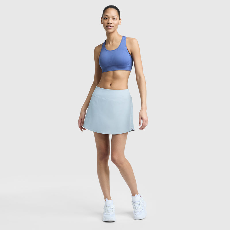 A woman stands against a plain light gray background, wearing the Champion Infinity Racerback Sports Bra, a light blue athletic skirt, and white sneakers. She smiles slightly with one hand on her hip.