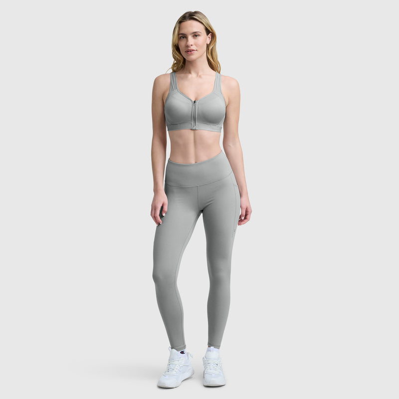 A woman stands against a plain light gray background, wearing the Champion Motion Control Zip Sports Bra, matching high-waisted leggings, and white athletic shoes. She faces forward with a neutral expression.
