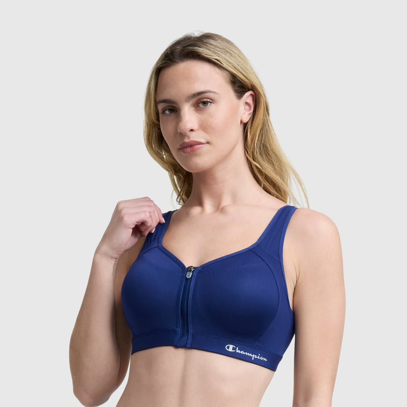 A woman with long blonde hair wears a blue Champion Motion Control Zip Sports Bra with the logo on the band, standing against a light gray background.