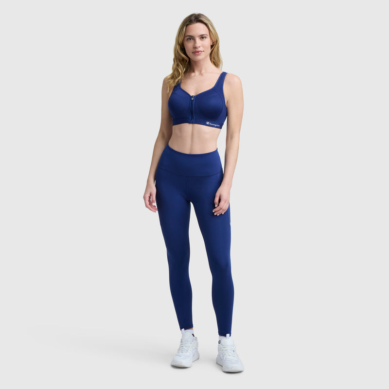 A woman stands against a plain light background, wearing the Champion Motion Control Zip Sports Bra in navy with matching high-waisted leggings and white sneakers. She has long, wavy hair and looks forward with a neutral expression.