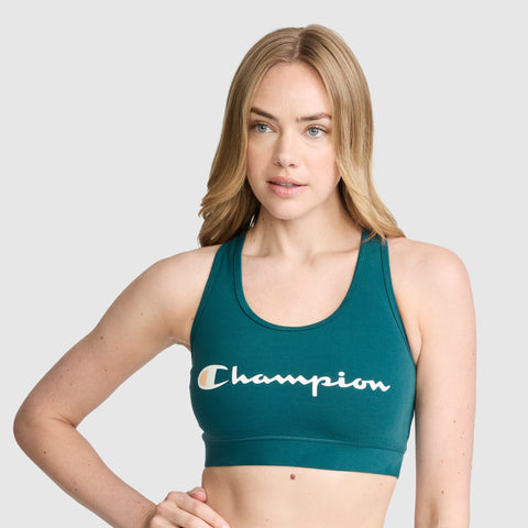 A woman with long blonde hair models the Champion Sports Bra, Script Logo—an athletic bra made from a cotton-spandex blend—in teal, standing against a plain light gray background.