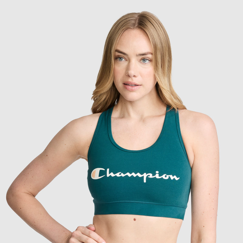 A woman with long blonde hair models the Champion Sports Bra, Script Logo—an athletic bra made from a cotton-spandex blend—in teal, standing against a plain light gray background.