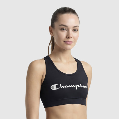 A woman with straight brown hair tied back is wearing a black Champion Sports Bra, Script Logo. Made from soft cotton-spandex jersey, she stands against a plain light gray background, smiling softly.