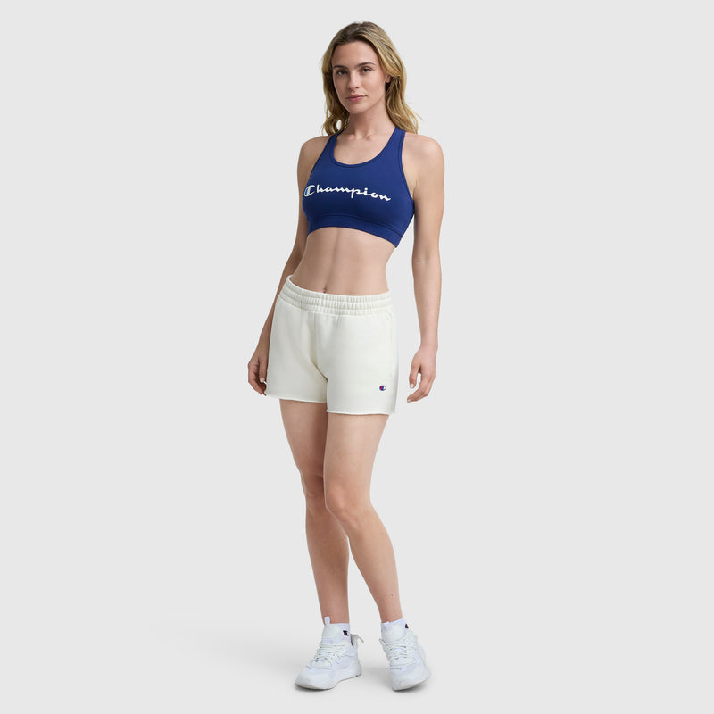 A woman faces forward wearing a navy Champion Sports Bra with Script Logo by Champion, cream shorts, and white sneakers, set against a plain light gray background.