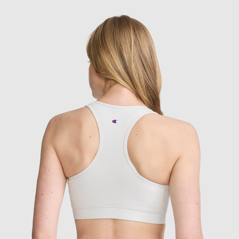 A woman with long blonde hair is shown from behind against a plain light background, wearing the Champion Sports Bra, Script Logo—a white cotton-spandex athletic bra with a small Champion logo on the back.
