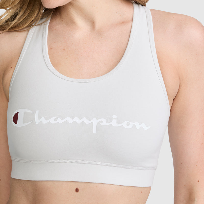 A person is shown from the shoulders to the waist on a plain background, wearing a light gray Champion Sports Bra, Script Logo. This athletic bra displays the classic Champion script logo across the front for a sporty look.