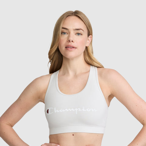 A woman with long blonde hair is wearing the Champion Sports Bra, Script Logo—a light gray cotton-spandex athletic bra featuring “Champion” in white—while standing with her hands on her hips against a plain light background.