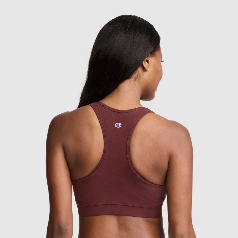A woman with long dark hair is seen from the back, wearing a maroon Champion Sports Bra, Script Logo. This bra features a classic racerback design and is made from a soft cotton-spandex blend for comfort. The Champion logo appears at the center of the upper back. The background is plain and light gray.