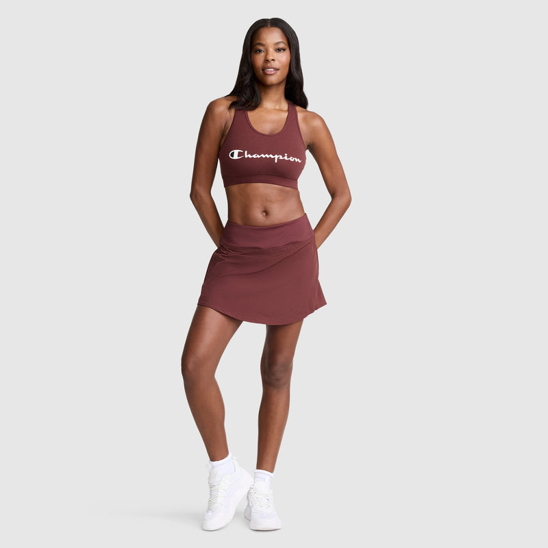 A woman wearing a maroon Champion Sports Bra with a script logo and a matching athletic skirt, paired with white sneakers, stands with one hand on her hip and smiles against a plain light background.