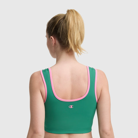 A woman with blonde hair in a ponytail is shown from the back, wearing the Champion Courtside Seamed Bra Top in green with pink trim and a small logo. The plain light gray background highlights her athletic style.