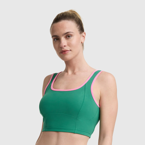 A woman with light skin and blonde hair in a ponytail wears the Champion Courtside Seamed Bra Top, a green performance fabric sports bra with pink trim, standing against a plain light gray background and facing forward.