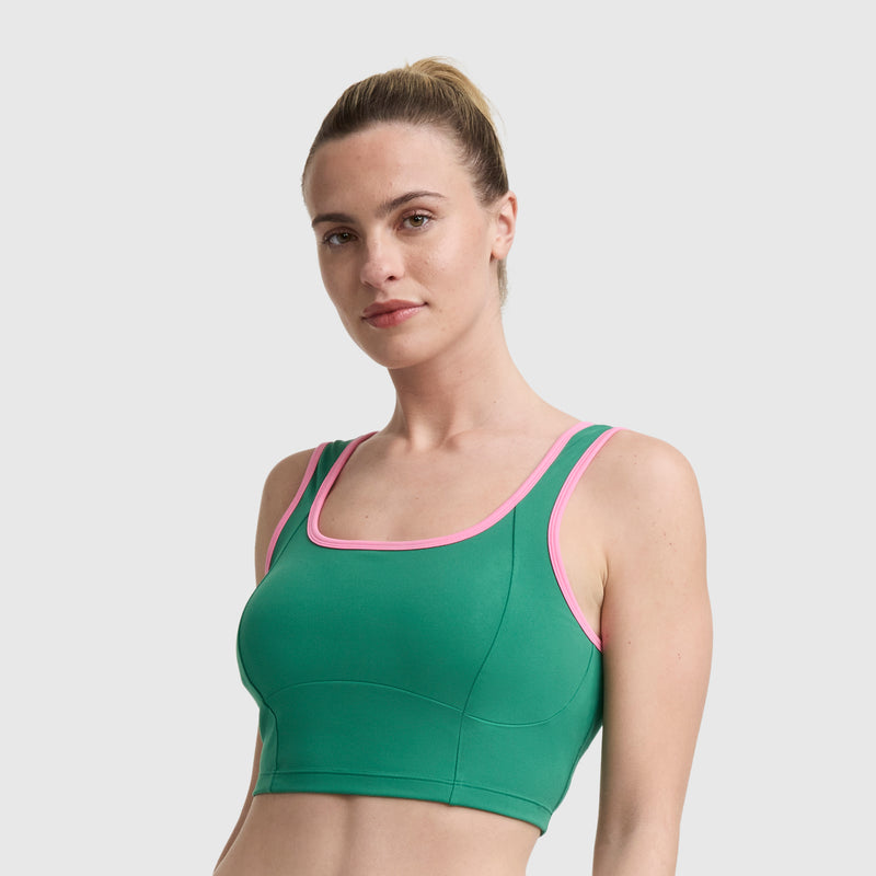 A woman with light skin and blonde hair in a ponytail wears the Champion Courtside Seamed Bra Top, a green performance fabric sports bra with pink trim, standing against a plain light gray background and facing forward.