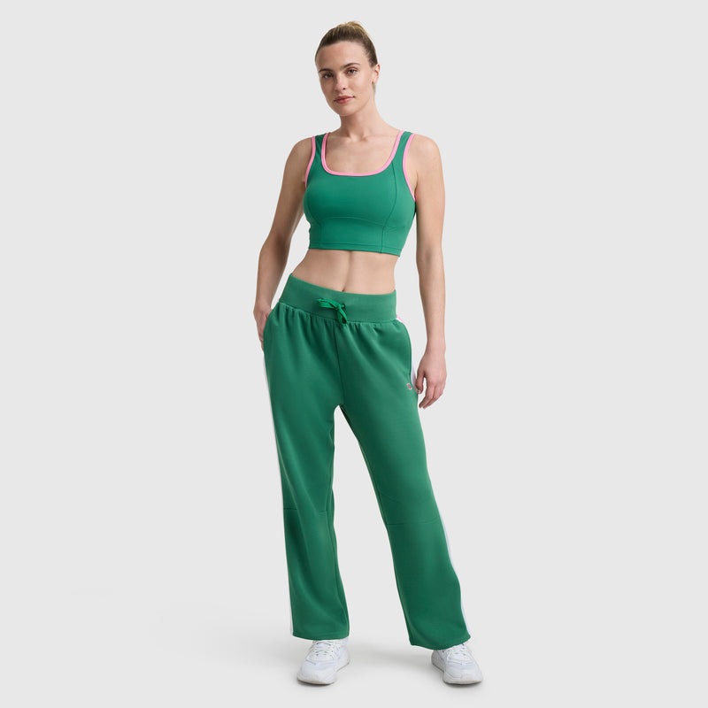 A woman with her hair pulled back models the Champion Courtside Seamed Bra Top in green with pink trim, paired with green drawstring pants and white sneakers against a white background, creating a sporty, athletic look.