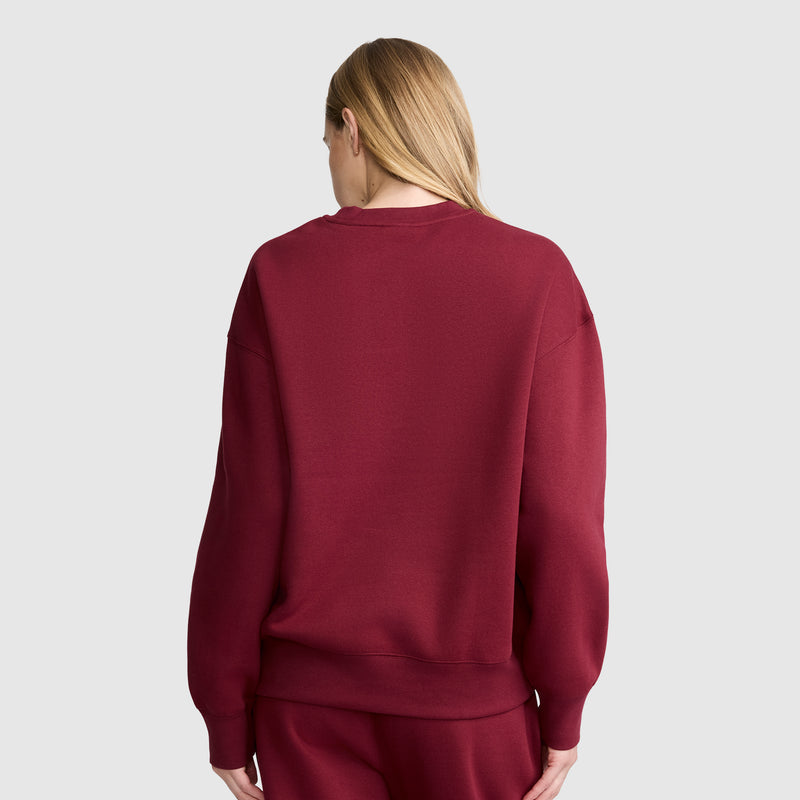 A person with long blonde hair is standing with their back to the camera, wearing a maroon Champion Powerblend Crewneck Sweatshirt, Script Logo, paired with matching pants made from soft fleece, set against a plain light gray background.
