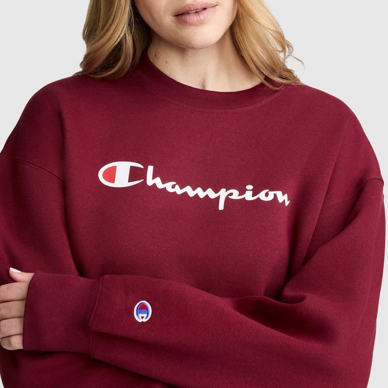 A person wearing the Champion Powerblend Crewneck Sweatshirt, Script Logo, in maroon. The sweatshirt features the iconic brand logo across the chest and a small Champion patch on the sleeve. Its classic crewneck design is crafted from cozy Powerblend fleece for optimal comfort and warmth.