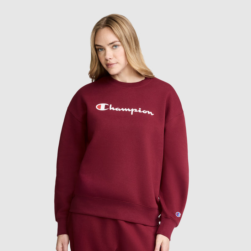 A woman stands against a plain light gray background, looking slightly to the side with a neutral expression. She is wearing the Champion Powerblend Crewneck Sweatshirt, Script Logo in maroon with matching pants—a classic crewneck crafted from soft Powerblend fleece for comfort and style.