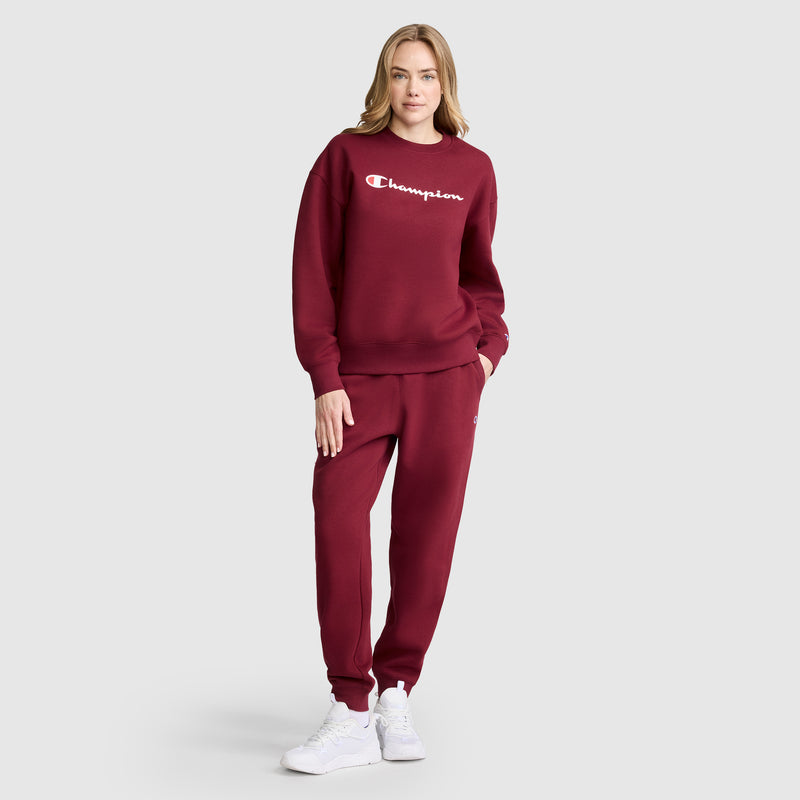 A woman stands wearing the Champion Powerblend Crewneck Sweatshirt with Script Logo in maroon, paired with matching sweatpants and white sneakers. One hand rests in her pocket as she looks at the camera against a plain gray background.
