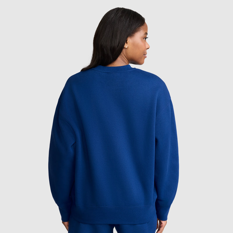 A woman stands facing away from the camera, wearing a bright blue Champion Powerblend Crewneck Sweatshirt with a script logo and matching pants against a plain light gray background.