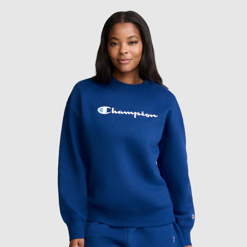 A woman wearing the Champion Powerblend Crewneck Sweatshirt, Script Logo, in blue stands against a plain light gray background, looking at the camera with a slight smile. The classic crewneck style and Powerblend fleece fabric from Champion combine comfort and timeless appeal.