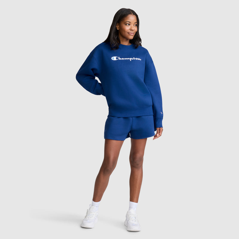 A woman wearing a Champion Powerblend Crewneck Sweatshirt, Script Logo in blue and matching shorts stands smiling with one hand on her hip, posing against a plain light gray background. Crafted from soft Powerblend fleece, the classic crewneck adds a cozy yet stylish touch. She completes the look with white sneakers and small hoop earrings.