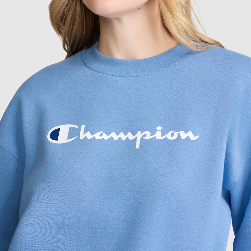 A person is wearing a light blue Champion Powerblend Crewneck Sweatshirt, Script Logo, featuring the Champion brand name printed in white across the chest. The cozy fleece sweatshirt adds comfort, while the person’s face is partially out of frame.